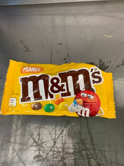 A yellow package of M&M's Peanut chocolate candies is displayed on a scratched, dark gray metallic surface. The package features the M&M's logo prominently with the words "PEANUT" in an orange oval above it. Nutritional information is visible on the left side of the package. A cartoon M&M character, red with white gloves and shoes, is depicted holding a single M&M. The image was taken in Worms, Germany, but no specific location within the city is identifiable from the visual evidence.