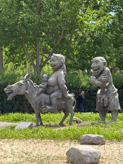 A park scene in Changping District, China, exhibits two prominent sculptures. One features a figure riding a donkey, accompanied by what appears to be a child. The other sculpture portrays a standing figure, seemingly clutching its arms to its chest. Between them, a man is visible, standing and holding a mobile phone. The surrounding area consists of trees, grass, and scattered rocks, suggesting a garden setting.
