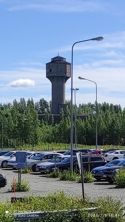 In Otanmäki, Finland, a concrete water tower with a hexagonal base and a cylindrical top stands prominently against a bright blue sky with scattered white clouds. The tower, featuring visible windows and a visible metal railing at its apex, is surrounded by a dense forest of evergreen trees. In the foreground, a gravel parking lot is filled with several cars, including a white SUV, a dark-colored sedan, and a black van. Two tall streetlights with modern, curved designs are positioned in front of the tower, adding to the urban infrastructure visible in the otherwise natural landscape. The scene is captured on a clear day, with the sun casting shadows and highlighting the textures of the environment.