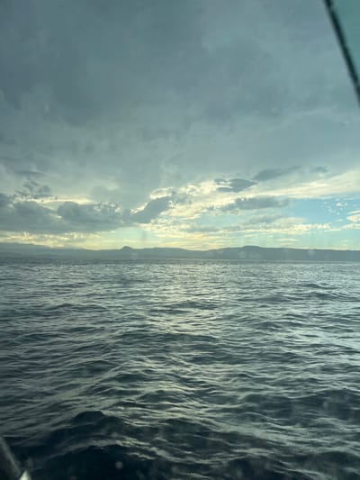 The image shows an ocean view with a horizon line running horizontally across the center. Dark, rippled water occupies the right half. On the left, there is a blend of dark and light clouds, with a section of light, partly obscured sky near the horizon. A landmass appears as a thin, darker band along the horizon, separating the sky from the water. The overall scene conveys a sense of movement and a potentially stormy atmosphere.
