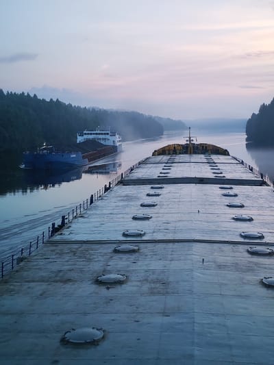 This is an early morning or late evening aerial view of a river in Volokov Most, Russia, with two barges. The sky is a pale blue and pink gradient, with some clouds. A dense forest lines both sides of the river. In the foreground, the deck of a large barge is visible, with numerous circular hatches. In the middle ground, a blue barge is heading towards the viewer, loaded with dark material, possibly coal, and is being pushed by a white vessel with the name "БАЛЫМ" visible on its hull. The water is calm, reflecting the sky and the barges.