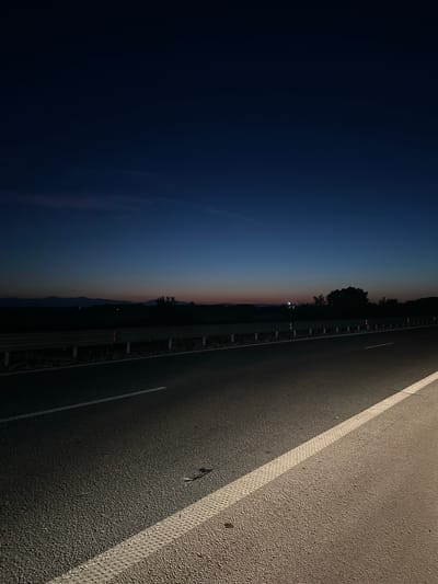 This image depicts a highway at dusk, with the sky transitioning from deep blue to lighter hues near the horizon. In the distance, a silhouetted landscape with mountains and trees is visible. The highway itself is paved, with a white line marking the lane. A guardrail runs along the side of the road, separating it from the adjacent terrain. There are no people or vehicles present in the scene. The overall atmosphere is one of tranquility and solitude.