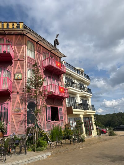 This image, taken in Măng Đen, Vietnam, captures a unique, multi-story building with a whimsical architectural style under a partly cloudy sky. The prominent structure in the foreground features a reddish-pink wooden facade composed of irregularly arranged planks and cross-braces, giving it a rustic yet playful appearance.

The building displays a variety of window shapes, including arched, round, and rectangular ones, some fitted with blinds. Several distinct pink-colored, box-like balconies protrude from the facade, two of which prominently display the national flag of Vietnam (red with a yellow star). The roof, which appears to be dark, possibly metal or tiled, is adorned with a decorative weather vane or sculpture on its peak. A partially obscured golden sign, seemingly starting with the letter "B", is visible on the left side of the roofline.

Adjacent to this distinctive building, to its right, stands a more conventionally designed multi-story structure, painted in a light cream or yellow color, featuring modern balconies with black wrought-iron railings.

The ground level in front of the pink building includes a lush garden area with various plants, shrubs, and flowering bushes, including yellow and purple blooms. Several dark metal outdoor tables and chairs are arranged on a paved brick or cobblestone surface, suggesting an outdoor seating area. A small, coniferous tree grows directly in front of the pink building.

In the background, a verdant, tree-covered mountain or hill rises, indicating a natural landscape surrounding the development. A dark-colored SUV and a red car are parked further down the paved road. The sky above is partly cloudy, with patches of bright blue visible amidst grey clouds, suggesting daytime with fluctuating sunlight.