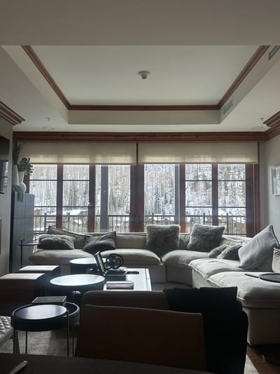 A living room features a large, neutral-toned sectional sofa adorned with plush pillows. An abstract sculpture sits on a dark coffee table. Floor-to-ceiling windows provide a snowy, outdoor view. Light-filtering shades partially cover the windows. Wooden trim accents the ceiling and window frames. A white entertainment center and a few plants add detail to the space. Two small tables are visible. A minimalist aesthetic pervades.