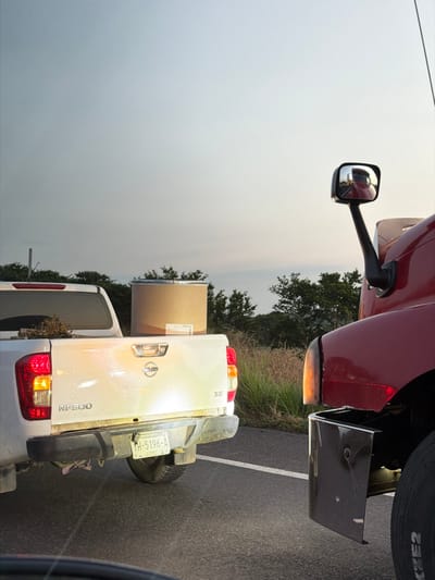 A white Nissan truck carries a large cardboard cylinder in its bed, partially obscured by a bright glare. The truck has a license plate and is equipped with a bumper. A red semi-truck with a side mirror is positioned beside it. The background includes sparse trees and what appears to be a hazy sky. The scene is set on a paved road.