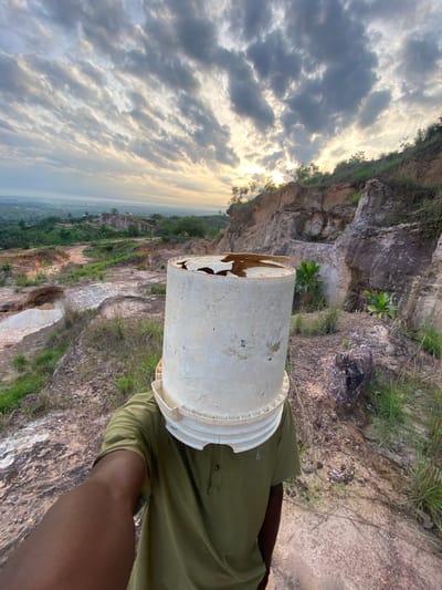 The image, oriented vertically, depicts a person standing on a rocky hillside or quarry in Ufueseni, Nigeria. The central subject is an individual whose head is obscured by a worn, white plastic bucket. The bucket shows signs of age and has a distinct damaged or torn section on one side. The person is wearing a short-sleeved, olive-green polo shirt, and their dark-skinned arm is visible below the bucket.
The background reveals a rugged, natural landscape dominated by large, exposed rock formations and cliffs, characteristic of a quarry or mountainous terrain. Patches of green grass and small plants are interspersed among the rocks. Beyond the immediate foreground, a vast expanse stretches out to the horizon, showcasing a valley or distant settlement under a dramatic sky. The sky is largely covered by white and grey clouds, with the sun partially breaking through, creating a soft, diffuse light that suggests either early morning or late afternoon. No specific activity is clearly depicted beyond the person posing with the bucket on their head, possibly for an artistic or humorous effect. There is no visible text in the image.