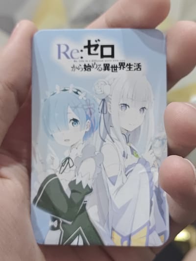 The image shows a close-up of a hand holding a rectangular anime-style keychain or card. The keychain features an illustration of two female anime characters, one with blue hair and the other with white hair. Both characters are wearing ornate outfits. The background of the keychain is a light blue with a subtle wave pattern. The image is taken from a slightly elevated angle, with fingers partially obscuring the edges of the keychain. The lighting suggests an indoor environment. There is no discernible location in the city of Bugulma, Russia.
