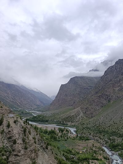 The image shows a mountainous valley with a river flowing through it. The sky is overcast with clouds. The slopes of the mountains are covered with sparse vegetation and rocky outcrops. In the distance, a small village is visible nestled in the valley. The village appears to have a cluster of white tents and some low-lying buildings. A road winds along the side of the valley. The image appears to be taken from a high vantage point, looking down into the valley. The location is specified as Gemur, India.