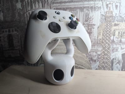 The image shows a white Xbox Series S/X controller resting on a white, 3D-printed stand resembling the Hollow Knight character. The controller appears dusty and used. The stand has two large, circular holes representing the character's eyes. The background is a wallpaper depicting various European landmarks, including what appear to be the Colosseum in Rome and the Eiffel Tower in Paris. The objects are placed on a light-colored wooden surface. The scene is not clearly indicative of being specifically located in Жаңа Қуат, Kazakhstan.