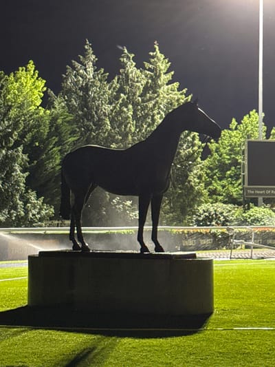 The image shows a statue of a horse at Kempton Park Racecourse in the Borough of Spelthorne, United Kingdom, taken at night. The dark statue is mounted on a cylindrical base and is positioned in front of trees. A scoreboard is visible behind the statue, partially obscured by the trees. Sprinklers are watering the green grass surrounding the statue and base. A barrier with "Unibet" printed on it is also visible in the background. The sky is dark, suggesting it is nighttime.
