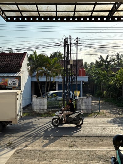 In Kedawung, Indonesia, a man wearing a black t-shirt and tan pants rides a white scooter. He is seen from the side, moving from left to right across the frame. The scooter is casting a long shadow on the asphalt road. In the background, a white truck is parked on the left, and a house with a red tile roof is visible on the left side of the image. There are palm trees and other foliage around the house, and utility poles with many wires crisscross the sky. The scene appears to be a street in a residential or commercial area, possibly with a shop called "TOKO ADIDA" indicated by a sign. The overall atmosphere is bright and sunny, with the light casting distinct shadows.