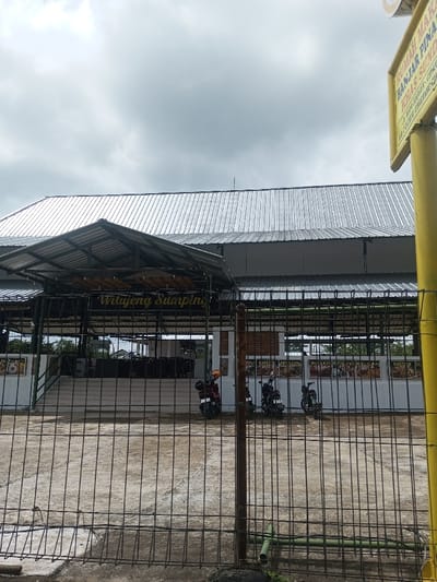 A low-angle view shows a building with a corrugated metal roof and open-air dining. A small awning covers the entrance with the words "Wilujeng Sumping." Three motorcycles are parked in front of a white wall adorned with food advertisements. A chain-link fence is in the foreground, and a yellow sign with Indonesian text is partially visible on the right. Overcast sky above.