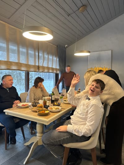 The scene depicts an indoor dining area, identified as being in Kalinovo, Russia. A group of individuals is gathered around a rectangular table, laden with various food dishes, wine bottles, and drinks.

In the foreground, a young male, likely a teenager, with short brown hair, is seated in a light-colored upholstered chair. He wears a white button-up shirt and a dark bow tie. He is actively waving his right hand towards the viewer with an open-mouthed smile. To his immediate left, a person, mostly obscured, is leaning over the table, wearing a light beige knitted sweater and dark pants.

Seated at the table on the left side are two adults. The man on the far left wears a dark long-sleeved top and has his hands clasped on the table. He looks towards the right. Next to him, a woman in a beige knitted sweater rests her chin on her left hand, looking towards the left.

In the mid-ground, standing behind the table, an adult male in a brown long-sleeved shirt is leaning against what appears to be a doorframe or wall, smiling. Further to the right in the background, another partially obscured individual is visible.

The table holds multiple plates with meals, including salads, a hamburger, and other prepared foods, along with a bottle of red wine, various glasses, and condiments. The setting features modern circular LED pendant lights suspended from a grey, slatted wooden ceiling. The background reveals large windows offering a view of a snowy, wooded landscape, suggesting a winter environment. Beige Roman blinds cover the upper portion of the windows. A small black security camera is mounted in the upper left corner near the ceiling. A large abstract painting with gold and grey tones hangs on the right wall. The floor is covered with dark, herringbone-patterned tiles.