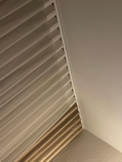 The image shows a section of a room's corner where the ceiling meets the wall, with curtains hanging from a track. The curtains are a light, semi-sheer fabric, and a darker, solid-colored curtain is visible behind the sheer one near the wall. The ceiling and wall appear to be painted in neutral tones. The image provides no indication of a specific location in Barutin, Bulgaria.