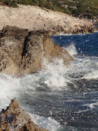 The image depicts a rocky coastline with waves crashing against the rocks. The water is a deep blue and appears choppy with white foam and spray. The rocks are jagged and vary in color from light beige to dark brown, with some appearing wet and glistening. Green vegetation grows on the hillside behind the rocks. The scene is illuminated by sunlight, creating highlights on the water and rocks. The location is identified as Karaburun, Turkey, suggesting a coastal area with natural rock formations. There are no people or man-made structures visible in the image. The focus is on the dynamic interaction between the water and the rocky landscape.