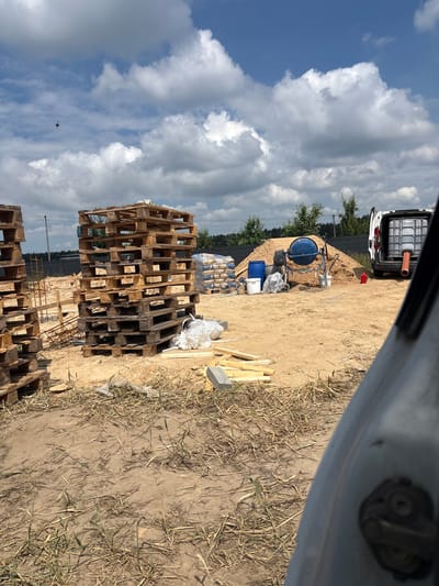 This image depicts a construction site located in Opatów, Poland. In the foreground, dry grass and dirt dominate the ground. Two stacks of wooden pallets are visible on the left. In the mid-ground, there is a large pile of sand with a blue cement mixer and various containers including white and blue buckets. A white van with a large translucent container and an orange pipe extending from it is parked on the right. Bags of cement are stacked behind the sand pile. The background features a dark fence and trees under a bright blue sky with scattered white clouds. A small dark object, possibly a bird or drone, is visible in the sky towards the upper left. The overall scene suggests an active construction or building project in an outdoor setting.