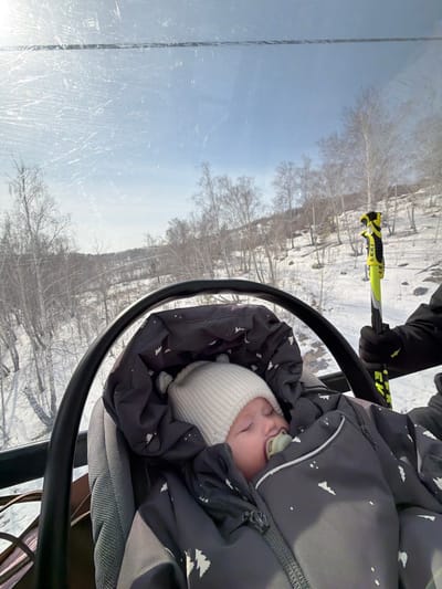 An infant sleeps securely in a dark gray, insulated car seat or stroller attachment, featuring a white tree pattern, inside an enclosed cabin, likely a gondola or ski lift, in Zelenaya Polyana, Russia. The infant wears a light-colored knitted beanie with small ear-like protrusions and has a pacifier in its mouth, with eyes closed. To the right, a black-gloved hand is visible, gripping a yellow ski pole branded "LEKI." The scene outside is viewed through a scratched transparent window, revealing a snow-covered winter landscape with numerous bare deciduous trees, primarily birches, scattered across a hillside under a clear blue sky.