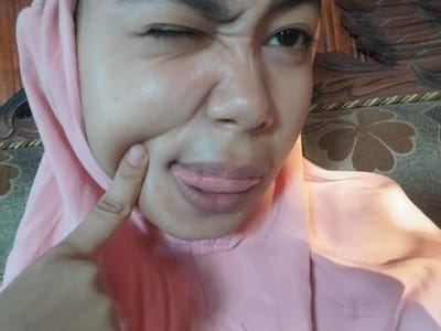 A close-up shot shows a woman's face from the chest up. She is wearing a pink hijab and a matching pink top. Her left eye is squinted shut, and her right eye is open, looking towards the camera. Her mouth is open, with her tongue sticking out. Her right index finger is touching her cheek. The background shows a textured wooden surface and a patterned cushion, suggesting an indoor setting, possibly in Labissa, Indonesia.