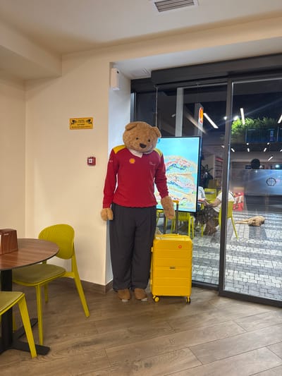 The image depicts an indoor scene, likely a cafe or rest stop, with a large teddy bear mascot standing prominently in the foreground. The mascot is dressed in a red long-sleeved shirt with yellow accents and a Shell logo, dark gray pants, and brown shoes. It is standing next to a bright yellow suitcase.
In the background, visible through a large glass window, is an outdoor area with cobblestone pavement. Several people are seated at yellow chairs, and a dog is lying on the ground. Inside, there is a round wooden table with a tissue dispenser and two yellow chairs with green cushions. On the wall, a yellow sign with an image of a CCTV camera and text in Armenian is visible, along with a red fire alarm box. A large screen displaying an image of stacked sandwiches is also present behind the mascot. The lighting suggests it is either evening or a dimly lit interior. Based on the context provided, this scene is located in Parakar, Armenia.