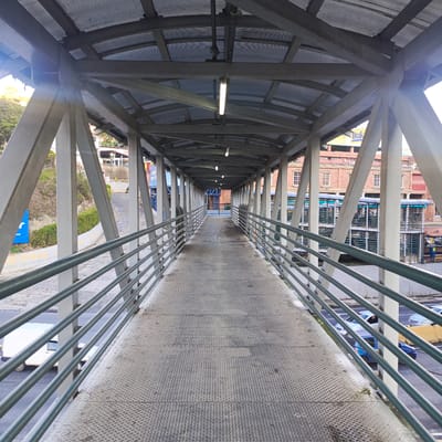 The image depicts a covered pedestrian overpass constructed with a metal frame and a corrugated metal roof. The walkway itself is made of diamond plate metal. Metal railings line both sides of the walkway. In the background, a multi-story brick building with white-framed windows is visible, along with what appears to be a parking structure or multi-level transportation hub. Several vehicles, including cars and a white van, are present on the road beneath the overpass. The environment suggests an urban setting, likely a transportation hub or a busy area within the city of Carrizal, Venezuela. The lighting suggests it is daytime, possibly overcast or late afternoon, given the shadows and the quality of light.