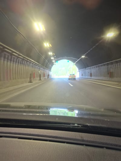 The image depicts the interior of a car driving through a tunnel, likely the tunnel on the road towards Vrachesh, Bulgaria, given the provided location context. The perspective is from the driver's seat, looking forward through the windshield. The tunnel is illuminated by overhead lights, casting a bright glow. The walls of the tunnel are visible on either side, with a ribbed texture. A white car is visible in the distance, driving in the same direction. The road surface appears to be asphalt, with lane markings visible. The foreground shows the dashboard of the car. The overall scene is one of travel through an urban infrastructure.