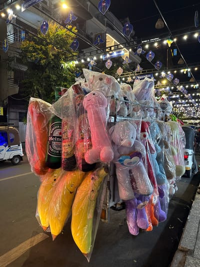 In Khan Daun Penh, Cambodia, at night, a street vendor's cart is laden with numerous soft toys and novelty items. The cart displays a variety of plush animals, including teddy bears in various colors such as light purple, gray, and a light blue one holding a rainbow heart. There are also phallic-shaped pink plush toys and what appear to be large, yellow banana-shaped plush toys with smiley faces. Interspersed among the toys are two green Heineken beer bottles and a bottle labeled "Kinh Kieu Ginseng". The entire cart is covered in clear plastic sheeting. In the background, string lights are strung across the street, adorned with decorative heart-shaped cutouts featuring figures. A small, white and blue tuk-tuk is parked to the left of the cart. The street is paved, and a concrete curb is visible. Buildings with lit windows line the street.