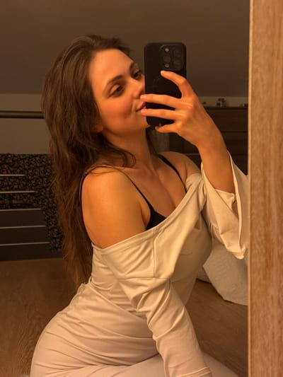 A female subject is depicted in a mirror selfie within an indoor environment. She is positioned facing a mirror, holding a black smartphone with a multi-lens camera array and a pop-socket in her right hand, partially obscuring the right side of her face. Her head is tilted slightly upward and to the right, with her gaze directed towards the phone's lens. Her lips are slightly pursed in a subtle smile. She has long, dark brown hair that falls over her left shoulder and down her back. She is wearing a light beige, long-sleeved garment that is pulled down to expose both shoulders, revealing a black spaghetti-strap top or bra underneath. The background, visible in the mirror's reflection, includes a dark, patterned fabric draped over horizontal bars to the left of the subject, and a dark wooden dresser with several small, light-colored objects on its surface to her right. The flooring in the lower foreground is composed of light-colored wooden planks. The lighting is warm and ambient. The specific location within Cava de' Tirreni, Italy, is not identifiable from the visual evidence presented in the image.