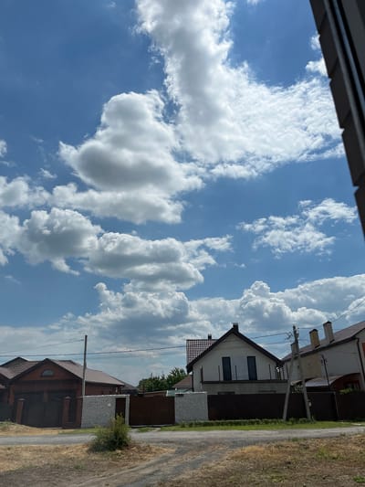 The image depicts a street view in Taganrog City District, Russia, under a partly cloudy blue sky. In the foreground, a dirt and gravel road runs horizontally. To the left and center, residential houses are visible behind fences. One house on the left has a dark red roof and dark green gates. The central house features a white facade with dark-paned windows and a dark roof. To the right, another house with a lighter facade and a reddish-brown roof is partially visible. Power lines and poles are present above the houses. The background is dominated by a bright blue sky filled with numerous fluffy white clouds.