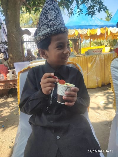 A young boy, dressed in a black long-sleeved shirt and traditional headwear, sits on a white chair. He holds a small cup filled with a dessert and a red spoon. He is smiling and looking to the right. In the background are trees, a blue and yellow tent, and other chairs. A woman wearing a hijab is partially visible behind the boy. The date "05/02/2026 14:31" is printed on the bottom right corner of the image.