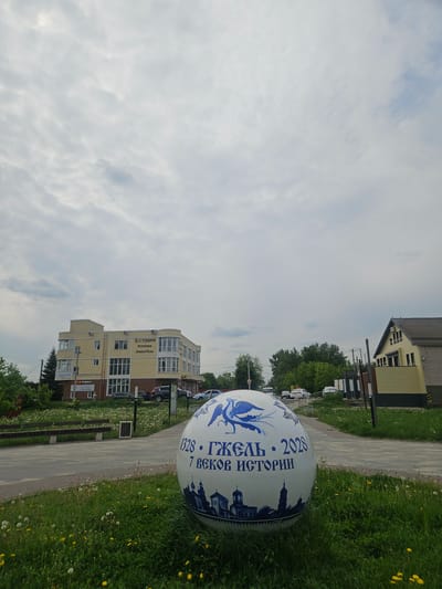 The photo shows an exterior scene in Речицы, Russia, featuring a large, spherical decorative object placed beside a paved walkway. The object is white with blue accents, depicting a stylized bird and the inscription "1328 • ГЖЕЛЬ • 2028, 7 ВЕКОВ ИСТОРИИ" in Russian.

The walkway is edged by grass, which contains dandelions. In the background, there are buildings, including a light-yellow building with the inscription "Студия Эстетики Лица и Тела" (Studio of Aesthetics for Face and Body). Also, there is a two-story house with light brown siding.

The sky is overcast with light clouds.