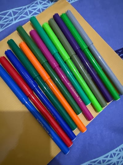 A close-up, angled shot shows a collection of multi-colored marker pens arranged diagonally on a golden-yellow surface, which appears to be a notebook or portfolio. The markers are predominantly in shades of green, purple, orange, red, and blue, with a few gray ones interspersed. The marker caps are visible, and some of the pens have "WATERCOLOR PEN" printed on them in white text. The golden surface rests on a blue fabric with a repeating geometric white pattern. The lighting suggests an indoor setting, possibly a desk or table. There are no people or discernible actions taking place, it is a still life composition of art supplies. The location of Pematang Siantar, Indonesia, is not identifiable from the visual information present in the image.