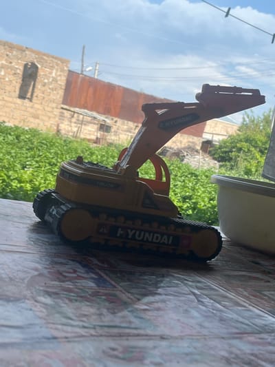 A toy excavator, yellow and orange, with "HYUNDAI" branding, sits on a patterned surface, possibly a tablecloth. The excavator is facing right, with its tracks and boom visible. In the background, out of focus, are green foliage, a brick building with an arched opening, and a rusted metal structure under a cloudy sky. The scene is viewed from a slightly elevated, close-up perspective. This image was taken in Jrarbi, Armenia.