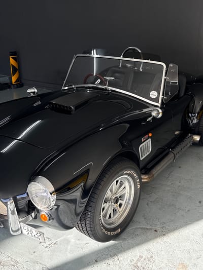 The image depicts a black convertible sports car, likely a replica of a Shelby Cobra, parked in an indoor garage with a dark grey wall in the background. The car is angled towards the right, showing its front-left side. It features a prominent hood scoop, a chrome windshield frame, side exhaust pipes, and a license plate reading "CAA 27 2164". The car has classic wire-spoke wheels with "BFGoodrich Radial T/A" tires. A yellow and black hazard bollard is visible to the left of the car. The floor is a light grey concrete surface with some dirt and shadows. No people are present in the image.