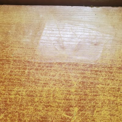 This is a close-up, top-down view of a worn wooden surface, possibly a table or countertop, in Ribina East, Nigeria. The wood is predominantly a reddish-brown color with a yellow, distressed finish applied in some areas, creating a mottled appearance. The surface is heavily scratched and shows signs of significant wear, with vertical grain lines visible beneath the damaged finish. A lighter, almost translucent patch is present on the left side, appearing to be a remnant of tape or some adhesive material that has partially peeled away, revealing a smoother, less scratched section of wood underneath. The overall texture is rough and aged, suggesting long-term use. There are no people or specific objects identifiable in the frame, only the textured surface itself. No text is visible. The lighting appears to be natural and diffused, casting no strong shadows, suggesting it could be daytime. The context of Ribina East, Nigeria, implies an outdoor or possibly indoor setting with tropical ambient conditions.