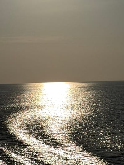The image displays an open body of water, likely the Aegean Sea, near Palaiopoli, Greece, under a hazy or overcast sky. The horizon line bisects the frame horizontally. The upper half is occupied by a muted grey-brown sky, exhibiting faint horizontal cloud bands. The lower half features the sea's surface, characterized by a prominent, intense reflection of sunlight forming an elongated, bright path extending from the horizon towards the foreground. This path is composed of numerous sparkling light points on the water, indicating surface ripples. The sun itself is not visible, but its intense glare dominates the water's reflection. No individuals, vessels, or other identifiable man-made objects are present.