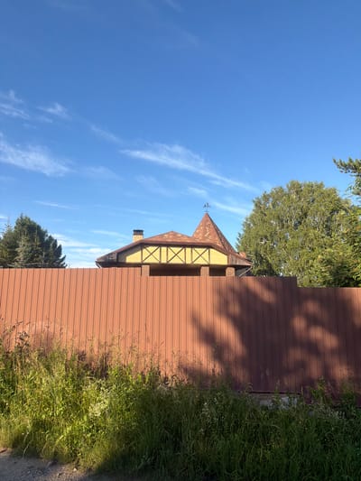 The image displays a sunny day in a rural setting. A brown metal fence runs horizontally across the right side of the frame. To the left of the fence, a portion of a building is visible. The building has a distinctive roof with brown, diamond-shaped shingles and yellow walls accented with dark trim. A brick chimney protrudes from the roof. A weather vane with a rooster silhouette sits atop the pointed roof. Green trees, including a large deciduous tree and a coniferous tree, are scattered throughout the scene, interspersed with the blue sky and wispy white clouds. The foreground on the right is bordered by tall, lush green grass with some wildflowers. The sun casts shadows from the trees onto the fence, indicating it is likely mid-morning or late afternoon. The overall impression is one of a peaceful, natural environment in what appears to be a village or countryside location, consistent with the provided location context of Колюткино, Russia. No people are visible in the image.