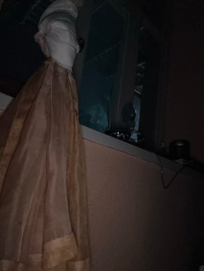 An indoor scene in Baygundi, Uzbekistan, captured under low light conditions, features a window area with associated objects and textiles. On the left, a vertically draped, light brown/tan, semi-translucent fabric, possibly a curtain or garment, hangs prominently. Positioned near the upper left of the window frame is a mannequin-like form, wrapped in white cloth with visible blue-grey banding around a head-shaped structure. The window, visible behind this form, reflects a dark blue-green hue suggesting an exterior. The window sill holds several items: a dark patterned bowl to the left, a small reflective spherical object, and a dark cylindrical container with a shiny lid to the right. A white cable extends across the sill from behind these objects. The wall on the right side of the frame is a muted reddish-brown or peach color, showing some texture. No individuals are present.