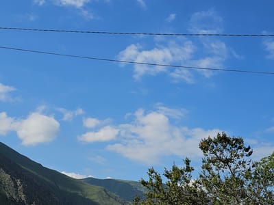 The image shows a clear blue sky with scattered white clouds. Two thin black wires run horizontally across the upper portion of the frame. In the lower right, the top branches of a green leafy tree are visible. In the lower left and extending towards the center, a steep, green mountainside dominates the landscape, with rocky outcrops and shadows cast by the sun. The scene appears to be a natural landscape, possibly on the outskirts of a city.