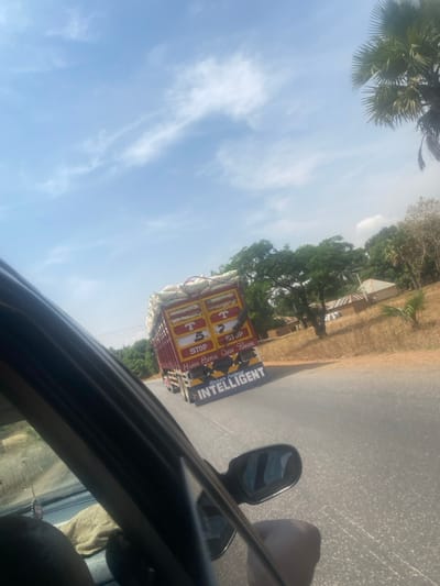 The image depicts a red cargo truck heavily laden with white sacks, traveling on a paved road in Bokana, Nigeria. The truck's rear is prominently decorated with yellow panels, black lettering, and red markings, including "STOP," the number "5," "HORN BEFORE OVERTAKING," "Baban Faduna," and "INTELLIGENT." The perspective is from the interior of a trailing or passing vehicle, likely a car, with a dark-skinned hand resting near the visible side window frame and mirror on the right. A light-colored cloth is visible inside the camera vehicle. The environment includes a bright blue sky with scattered white clouds, numerous green trees and dry foliage lining the road, and several small, light-colored structures with darker roofs in the distant background to the right. A large palm tree is visible on the far right.