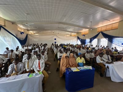 This image captures a large indoor gathering, likely an academic or educational event, in what appears to be a school hall or assembly room. Numerous young people, predominantly male students, are seated in neat rows at wooden desks. They are dressed in white long-sleeved shirts, with many wearing ties of various colors such as yellow, orange, red, and purple, paired with light brown or khaki trousers, consistent with school uniforms.

At the front of the room, on the left, a student is actively writing on papers placed on a white-covered table, which also holds green folders and books. On the right side, two women in light brown hijabs and long garments, alongside another woman in a yellow top, blue skirt, and blue head covering, are seated behind a table draped in blue fabric. This table also displays books and educational materials; visible text on one item includes "FINANCIAL" and "topic," with another book partially showing "Pillars of...".

The room is decorated with white sheer fabric draped artistically across the upper sections of the walls and windows, accentuated with swathes of dark blue fabric. Blue and white balloons, and blue fabric cut into star or cross shapes, are used as additional embellishments. The ceiling features a patterned white paneling, multiple ceiling fans (one clearly marked "ORIENTAL"), and fluorescent lighting. Natural light streams in through high windows equipped with horizontal blinds and protective metal bars, indicating a daytime event. The overall scene suggests an organized and attentive gathering, possibly a seminar, workshop, or competition.