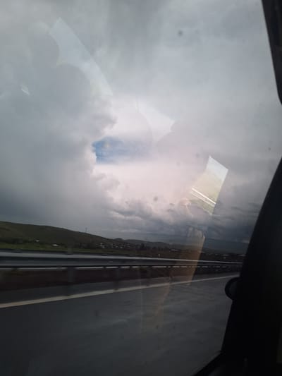 The image is a photograph taken from inside a vehicle, looking out through the window. It captures a cloudy sky over a rural landscape and a highway. The sky is dramatic, with dark storm clouds dominating the left side and lighter, brighter clouds on the right, suggesting an impending storm or the aftermath of one. There's a hint of blue sky visible through a break in the clouds.
In the middle ground, a green, rolling hillside is visible, dotted with small buildings and what appear to be some structures like cell towers or utility poles. A highway stretches horizontally across the frame in the foreground, with a guardrail separating it from the adjacent land. The road surface appears wet, indicating recent rain or humidity.
The right side of the image is partially obscured by the dark interior of the vehicle, likely the window frame and possibly a side mirror. There are reflections on the window glass, showing what appear to be the interior of the vehicle and possibly other elements from outside reflected in an unusual way. One prominent reflection shows a rectangular area with white lines, reminiscent of a runway or a marked surface.
The location is identified as Lusakert, Armenia. The scene depicts a common view along a highway in a semi-rural or mountainous region, characterized by its natural landscape and infrastructure. The overall mood is somewhat somber due to the cloudy weather.