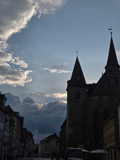 The image depicts a street scene in Ansbach, Germany, with a prominent church on the right. The church features two tall spires, one with a weather vane shaped like a rooster, and the other topped with a star. The church building appears to be made of stone and has gothic architectural elements, including arched windows. In the foreground, the street is lined with buildings of varying heights and architectural styles. On the left, multi-story buildings with light-colored facades display shop fronts, including one with an "H&M" sign. Further down the street, other buildings are visible, some with yellow facades and multiple windows.
The sky is partly cloudy with dramatic formations, and sunlight filters through some of the clouds, creating pockets of illumination. On the street, a few people are present, appearing as dark silhouettes due to the lighting conditions. A black van is parked in the lower right of the frame, and two white umbrellas are set up near it, suggesting an outdoor seating area. The overall atmosphere is one of a historic European city, with the imposing church dominating the skyline and the street life suggesting a typical urban environment.