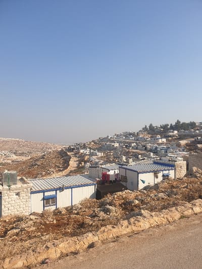 The correctly oriented image presents a wide view of a temporary settlement or camp situated within a dry, rocky, and hilly landscape under a vast, clear blue sky. The horizon appears hazy in the distance.
The foreground and mid-ground reveal numerous small, modular structures, predominantly white with blue roofs or trim, sprawling across the rugged terrain. These structures, likely prefabricated shelters, are densely packed in some areas and more scattered in others, adapting to the contours of the hillsides. A dusty, unpaved road runs along the upper right edge of the frame, bordered by a steep, rocky embankment covered with sparse, dry vegetation.
Detailing the foreground, several modular units are visible, some featuring windows. Laundry, including pink, red, and white clothing items, hangs on lines outside some of these shelters, indicating active habitation. Stone foundations or retaining walls are present beneath some units, suggesting basic site preparation. A dark-colored water tank is also discernible near one of the structures.
No people are visible in the image. The bright, clear sky suggests a sunny day, possibly late morning or early afternoon, with no clouds. The arid environment, coupled with the nature of the temporary housing, strongly points to an informal camp or an internally displaced persons (IDP) settlement. The location context of Killi, Syria, supports this interpretation. There is no visible text in the image.