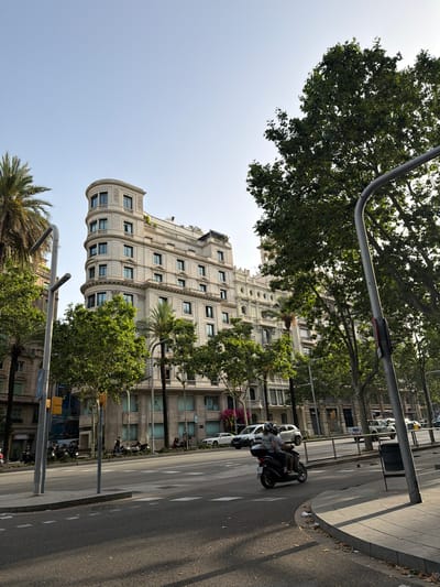 This is a street-level, eye-level, medium shot of a sunny day in Barcelona, Spain. The scene captures a curved building with light-colored stone facade and numerous windows on the left side, partially obscured by trees. In the foreground, a motorcycle with two occupants, wearing helmets, travels on the road. There are other vehicles and parked motorcycles visible further down the street, as well as trees lining the sidewalk and buildings in the background. The sky is clear and blue.