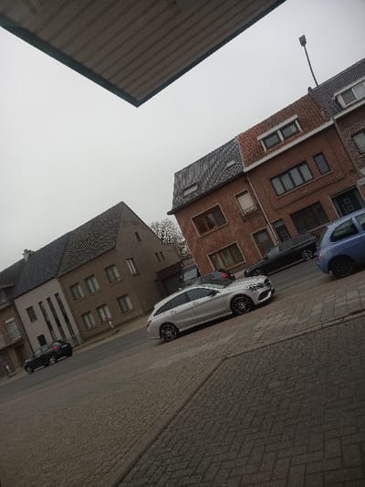 The scene depicts a street in Zwijndrecht, Belgium, observed under an overcast sky with a slight photographic tilt. A dark, corrugated metal overhang partially frames the upper portion of the image. The immediate foreground consists of a paved surface constructed from herringbone-patterned bricks.

On the right side of the road, a silver Mercedes-Benz C-Class Estate, featuring black multi-spoke wheels, is centrally positioned, facing right. A partial view of a light blue compact car is visible to its immediate right. Behind the Mercedes, a dark sedan is parked on the asphalt road. Further to the left, a dark-colored SUV is parked on the opposite side of the two-lane street.

Buildings line both sides of the thoroughfare. On the left, a light brown multi-story building with a dark grey tiled gable roof and several rectangular windows is visible. On the right, a red-brick multi-story building with a dark grey tiled roof, incorporating dormer windows and various window configurations, stands. A street light pole is discernible above the red-brick building. No individuals are present in the observable scene.