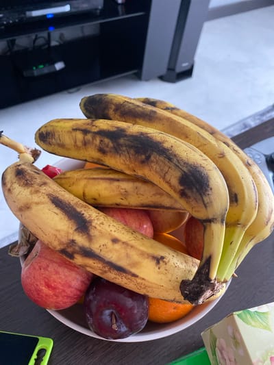 A close-up, eye-level shot shows a white bowl filled with various fruits, primarily ripe bananas with significant browning and some black spots. There are also a couple of red apples, a plum, and what appears to be an orange or mandarin. The fruits are arranged in a cluster, with the bananas dominating the composition and spilling out over the edge of the bowl. The bowl rests on a dark, possibly wooden, surface. In the background, out of focus, is a black electronic device and part of a shelf or furniture. The lighting is natural, creating shadows and highlighting the texture of the fruit. The scene appears to be indoors, likely a kitchen or living area. No people are visible in the image. The overall impression is of a still life featuring overripe fruit. This image is not located in Maksimkovo, Russia; it is a generic indoor scene.