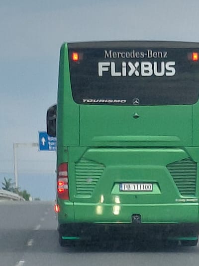 The image shows the rear of a green FlixBus coach with "Mercedes-Benz" and "FLIXBUS" branding. A license plate with the text "PO 111100" is visible. The bus appears to be on a road, possibly a highway, with a blue road sign showing an upward arrow and text in a foreign language visible to the left. The environment is overcast, with a hint of vegetation and a structure on the left side. The specific location in Voyvodinovo, Bulgaria is not directly discernible from the image content itself.
