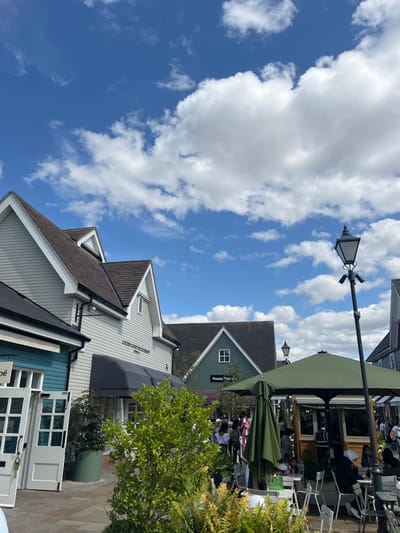 The image displays an outdoor retail environment under a bright blue sky with scattered white clouds. The location is identified as Bicester Village, situated within the Cherwell District of Oxfordshire, United Kingdom.
Architecturally, the scene features multiple buildings constructed in a village style. On the left foreground, a teal-colored building with open white double doors is partially visible, bearing a "Chloé" sign. Adjacent to it, a prominent light grey building with horizontal siding, a dark brown pitched roof, and multiple gables extends a black awning over its entrance. Above this awning, a sign reads "GOLDEN GOOSE DELUXE BRAND." In the mid-ground, a dark green building with a brown roof and a single white-framed window prominently displays "Vivienne Westwood" on its facade.
A paved pedestrian pathway extends throughout the scene. In the mid-right, an active outdoor café area is present, featuring numerous light-colored tables and chairs arranged beneath large, dark green octagonal umbrellas. A light-colored wooden kiosk is positioned under these umbrellas. Numerous individuals are visible; some are seated at the café tables, engaged in dining or conversation, while others are walking along the pathway. A tall, black, traditional-style lamppost with a lantern-like top stands in the right foreground. Green foliage, including a large bright green bush and ferns, is prominent in the foreground, complemented by darker green potted plants near the buildings. The scene is illuminated by strong natural sunlight.