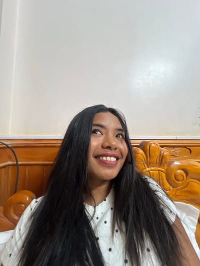 A medium shot shows a young woman with long, dark hair smiling and looking upwards in what appears to be a bedroom. She is wearing a white shirt with a black polka dot pattern. She is seated on a wooden bed frame that is intricately carved with ornate details. The background is a plain, light-colored wall. The lighting suggests an indoor setting, possibly during the daytime. The wooden furniture and the style of the bed might indicate a traditional Vietnamese aesthetic, however, without further information or context, it is not possible to pinpoint the exact location within Bắc Hà, Vietnam.