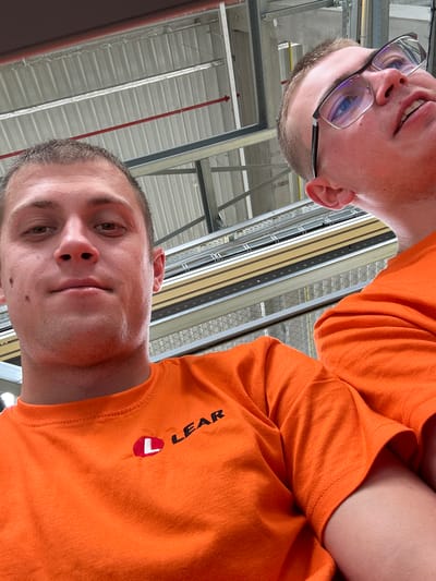 The image shows a close-up, upward-facing selfie of two individuals wearing orange t-shirts. The person on the left is wearing glasses with dark frames and has short brown hair. The person on the right is looking directly at the camera and has short brown hair and stubble. Both individuals appear to be in an industrial or warehouse setting, indicated by the visible metal structures and corrugated metal ceiling. The lighting suggests an indoor environment, possibly during the daytime. The t-shirt of the person on the right has a logo with the text "LEAR" and a red and white emblem. The overall mood is casual, with the subjects taking a selfie.